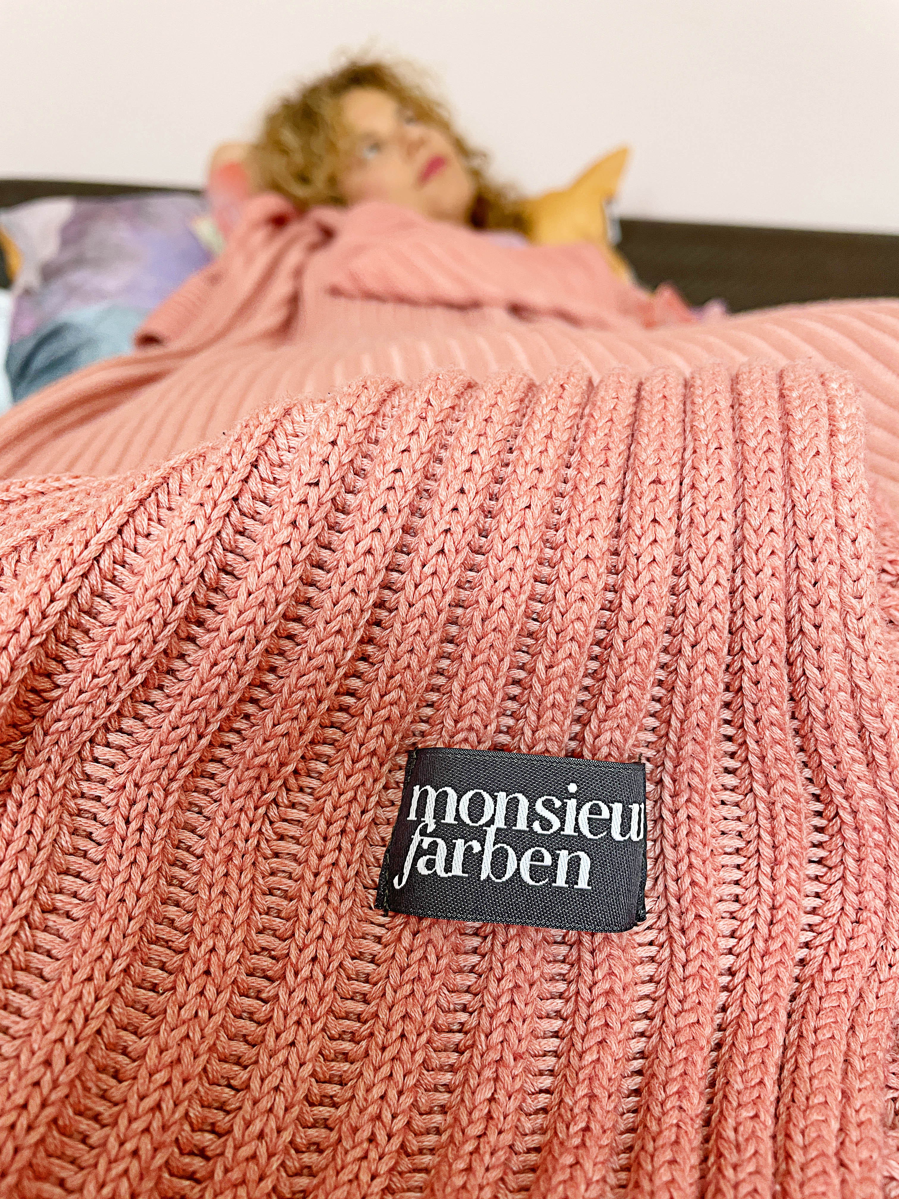 Eine Frau liegt verträumt im Bett. Auf ihr liegt eine gestrickte altrosa farbige Decke mit einem Label von der Marke Monsieurfarben.