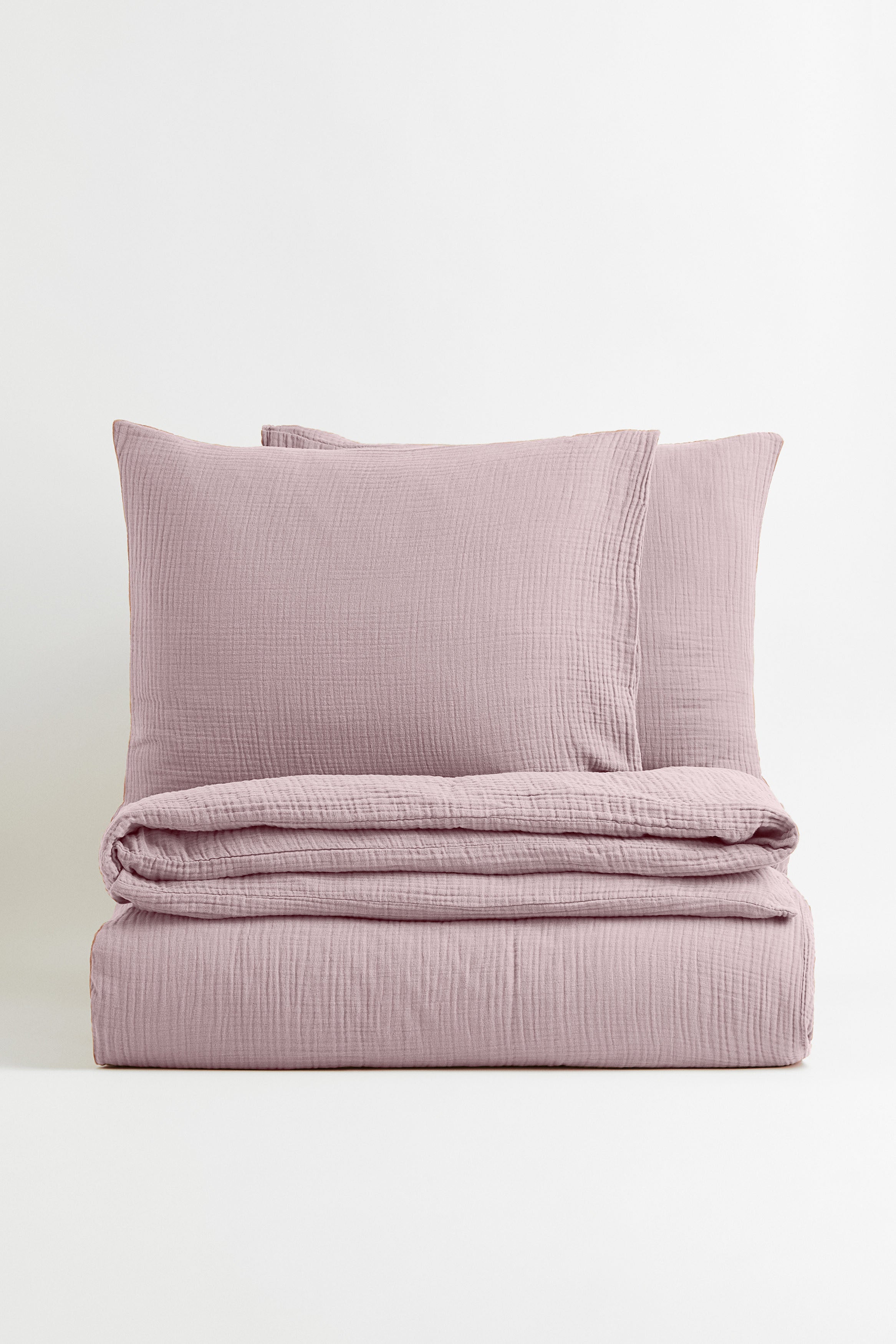 Auf dem Bild ist ein weißer Hintergrund mit einem Kissen und einer Bettwäsche drauf. Die Bettwäsche ist eine rosa Musslin Bettwäsche.