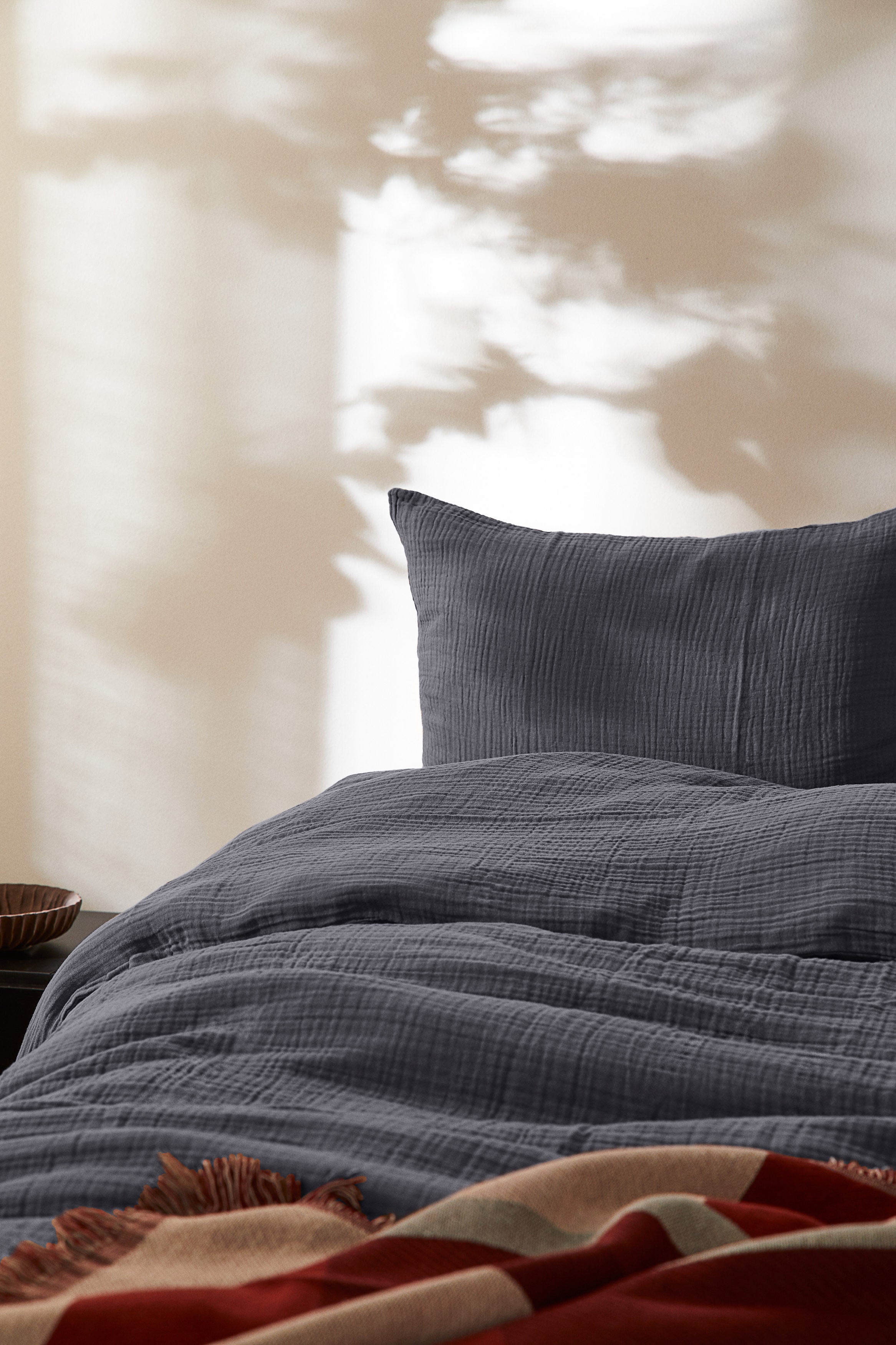 Auf dem Bild ist ein weißer Hintergrund mit einem Bett mit Kissen und einer Bettwäsche drauf. Die Bettwäsche ist eine graue Musslin Bettwäsche.