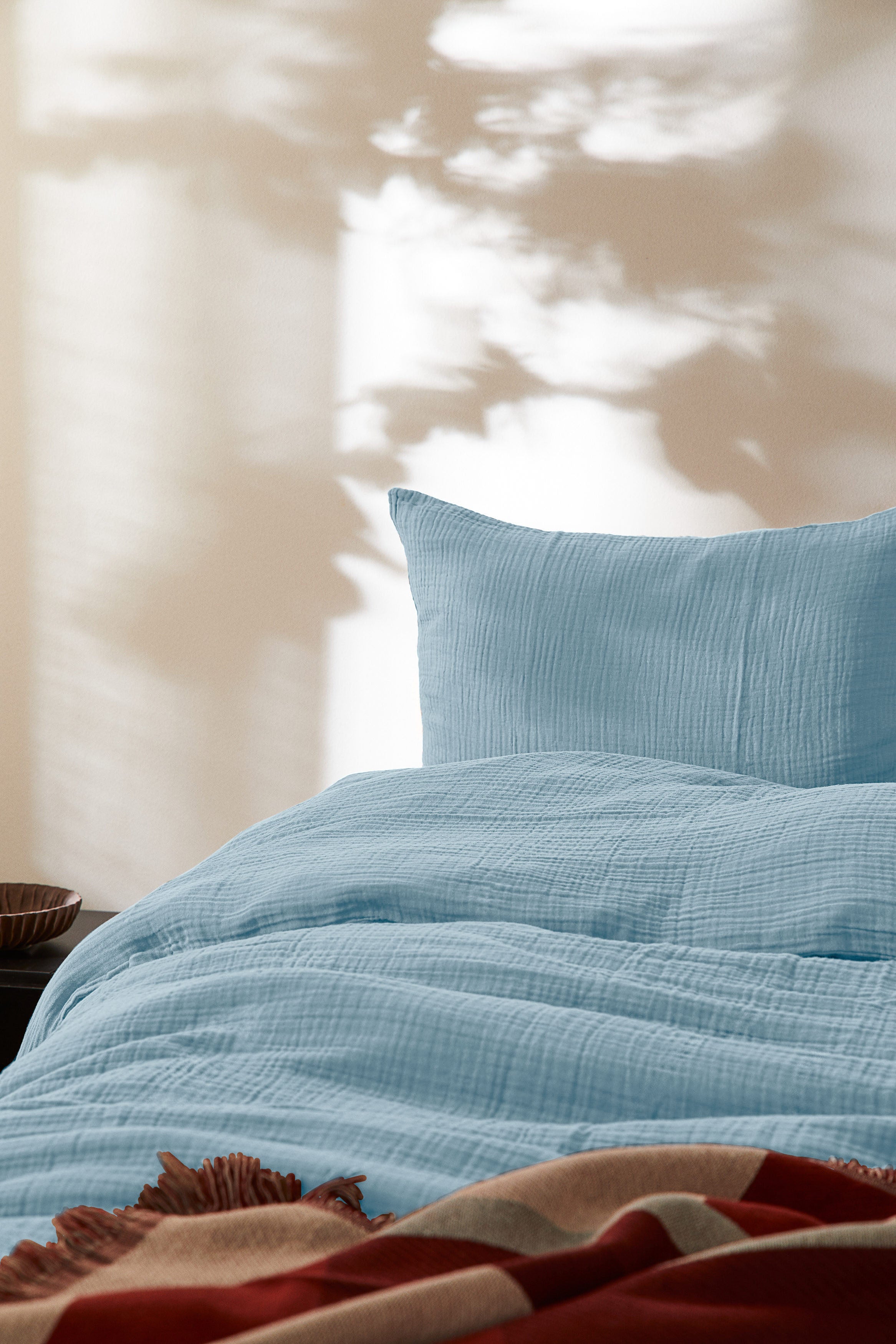 Auf dem Bild ist ein weißer Hintergrund mit einem Bett, Kissen und einer Bettwäsche drauf. Die Bettwäsche ist eine blaue Musslin Bettwäsche.