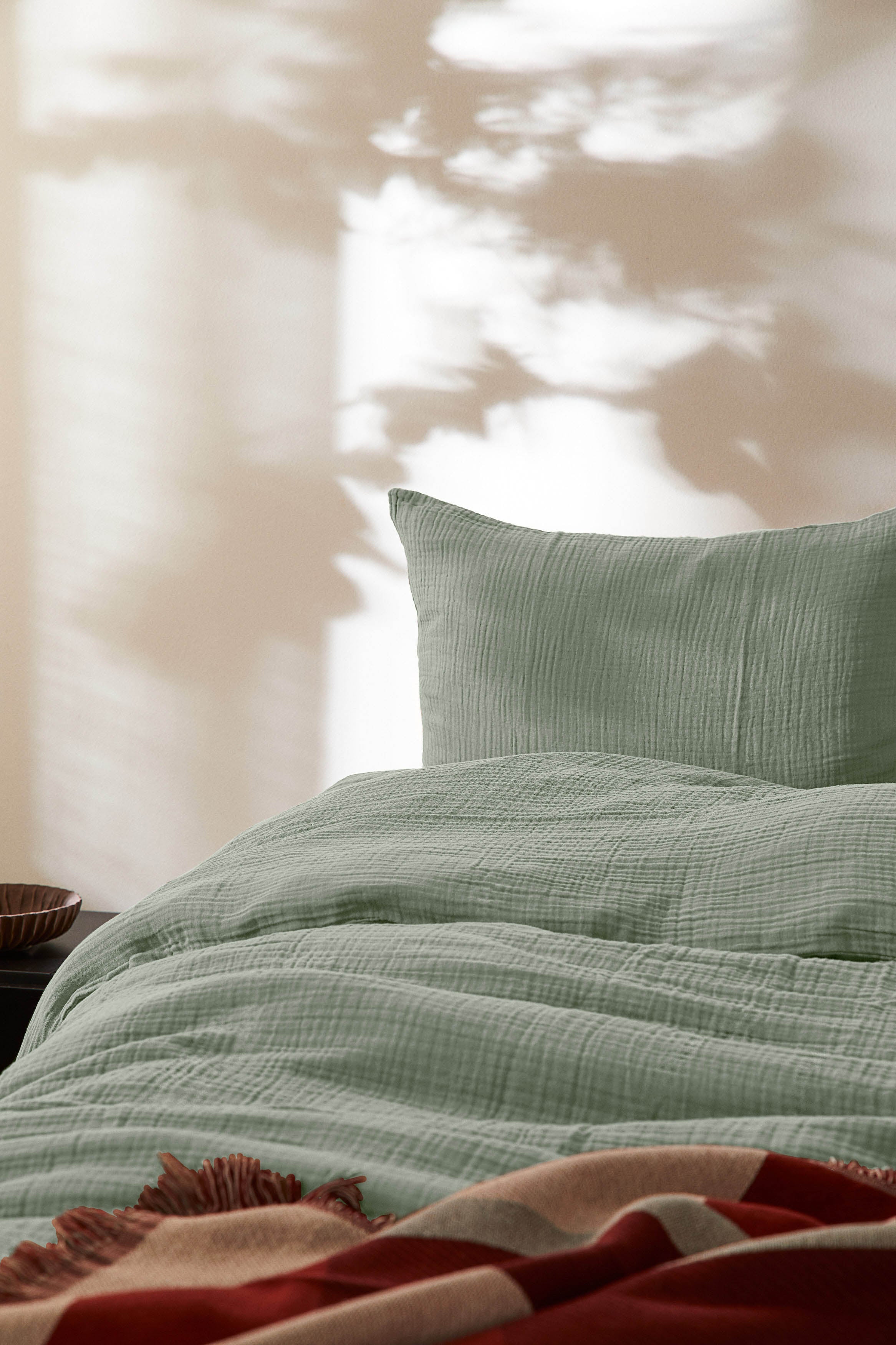 Auf dem Bild ist ein weißer Hintergrund mit einem Bett und einem Kissen und einer Bettwäsche drauf. Die Bettwäsche ist eine Grüne Musslin Bettwäsche.