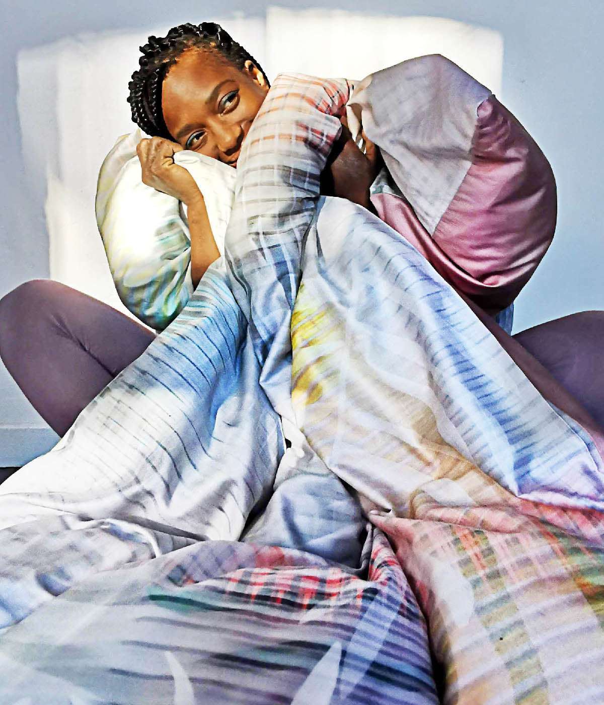 Dieses Bild zeigt eine Person, die sich gemütlich in eine Bettdecke mit einem tropischen Muster kuschelt. Die warmen Farben und das weiche Material verleihen dem Bild eine einladende, entspannte Atmosphäre. Das Licht im Hintergrund unterstreicht das Gefühl von Geborgenheit und Komfort.