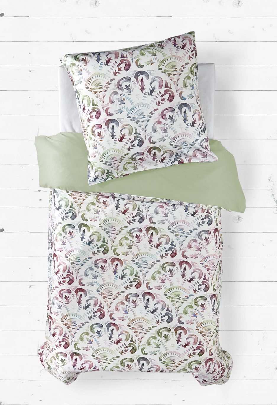 Eine Bettwäsche-Garnitur bestehend aus einem Bettbezug und einem Kissenbezug. Die Vorderseite zeigt ein detailliertes, symmetrisches Muster mit floralen und ornamentalen Elementen in Aquarelloptik, in Grün-, Blau-, Rot- und Lilatönen auf weißem Hintergrund. Die Rückseite ist in einem schlichten, pastellgrünen Farbton gehalten. Die Bettwäsche liegt flach ausgebreitet und wirkt hochwertig verarbeitet.