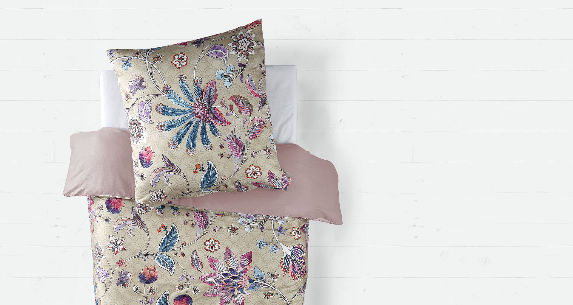 Ein Bett mit einer luxuriösen Bettwäsche im orientalischen Stil, die mit floralen Mustern in Blau-, Rosa- und Lilatönen auf einem beigen Hintergrund verziert ist. Das Kopfkissen trägt dasselbe Muster wie die Bettdecke. Die Bettwäsche wird durch ein schlichtes, altrosa Spannbettlaken ergänzt. Der Hintergrund besteht aus einer weißen Holzbodenoptik, die einen modernen und eleganten Kontrast zum opulenten Design der Bettwäsche bildet.