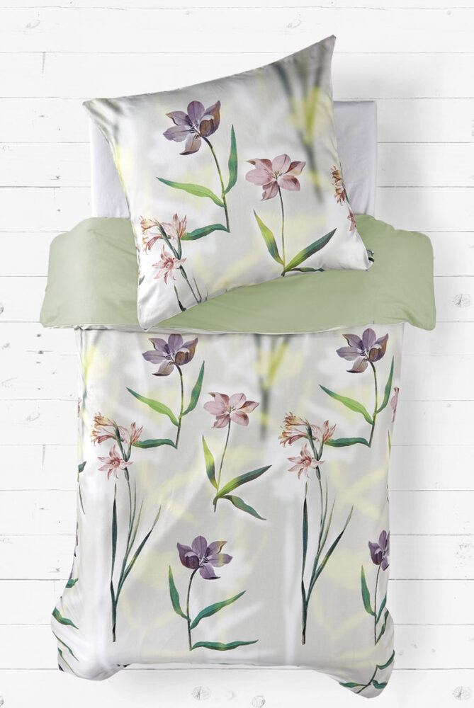 Bettwäsche-Set mit floralen Motiven auf hellem Hintergrund. Das Design zeigt violette, rosa und lachsfarbene Blumen mit grünen Stängeln und Blättern. Die Unterseite des Bettbezugs und das Laken sind in einem sanften Grünton gehalten. Das Bett liegt auf einem weißen Holzboden.