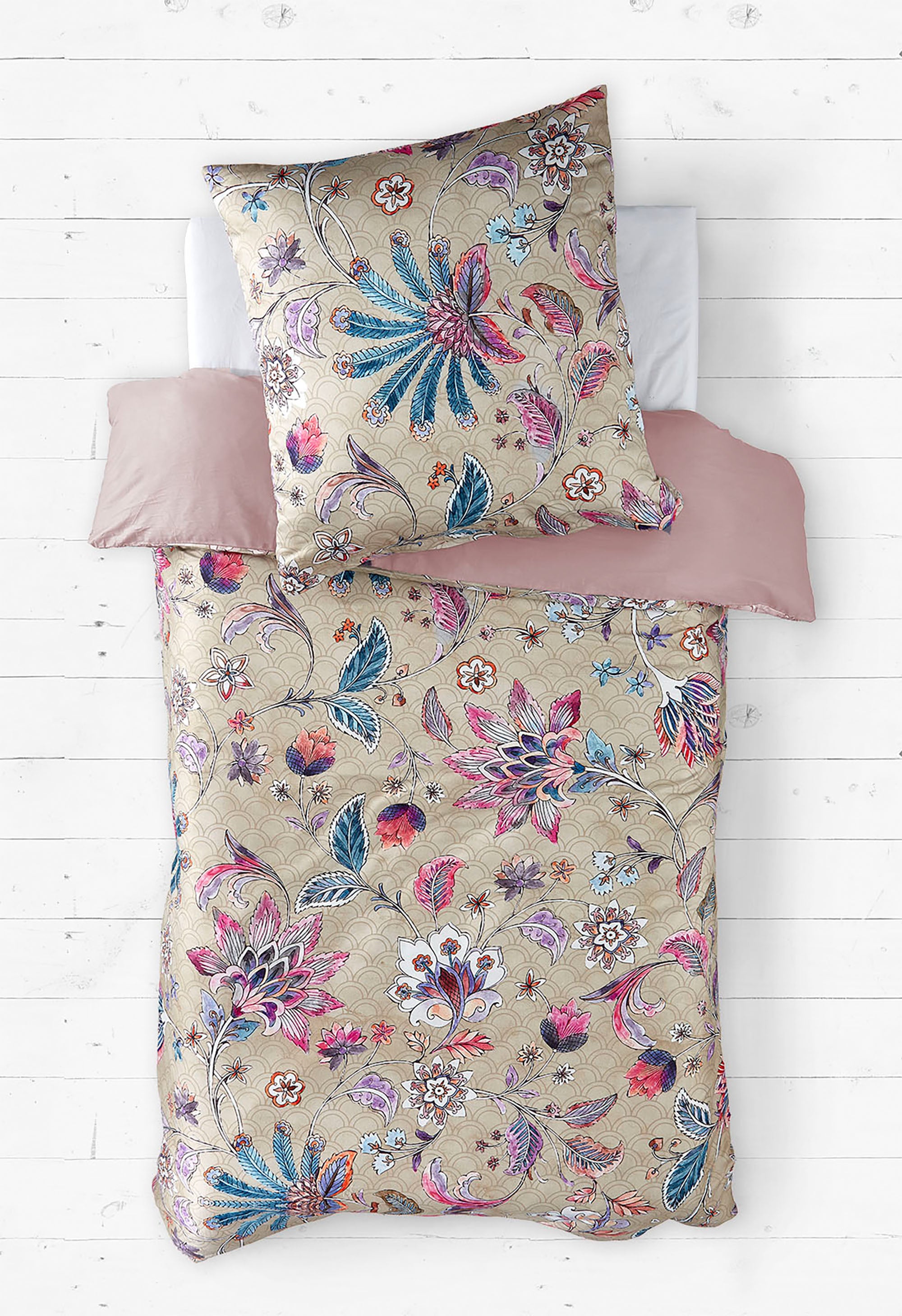 Ein Bett mit einer luxuriösen Bettwäsche im orientalischen Stil, die mit floralen Mustern in Blau-, Rosa- und Lilatönen auf einem beigen Hintergrund verziert ist. Das Kopfkissen trägt dasselbe Muster wie die Bettdecke. Die Bettwäsche wird durch ein schlichtes, altrosa Spannbettlaken ergänzt. Der Hintergrund besteht aus einer weißen Holzbodenoptik, die einen modernen und eleganten Kontrast zum opulenten Design der Bettwäsche bildet.