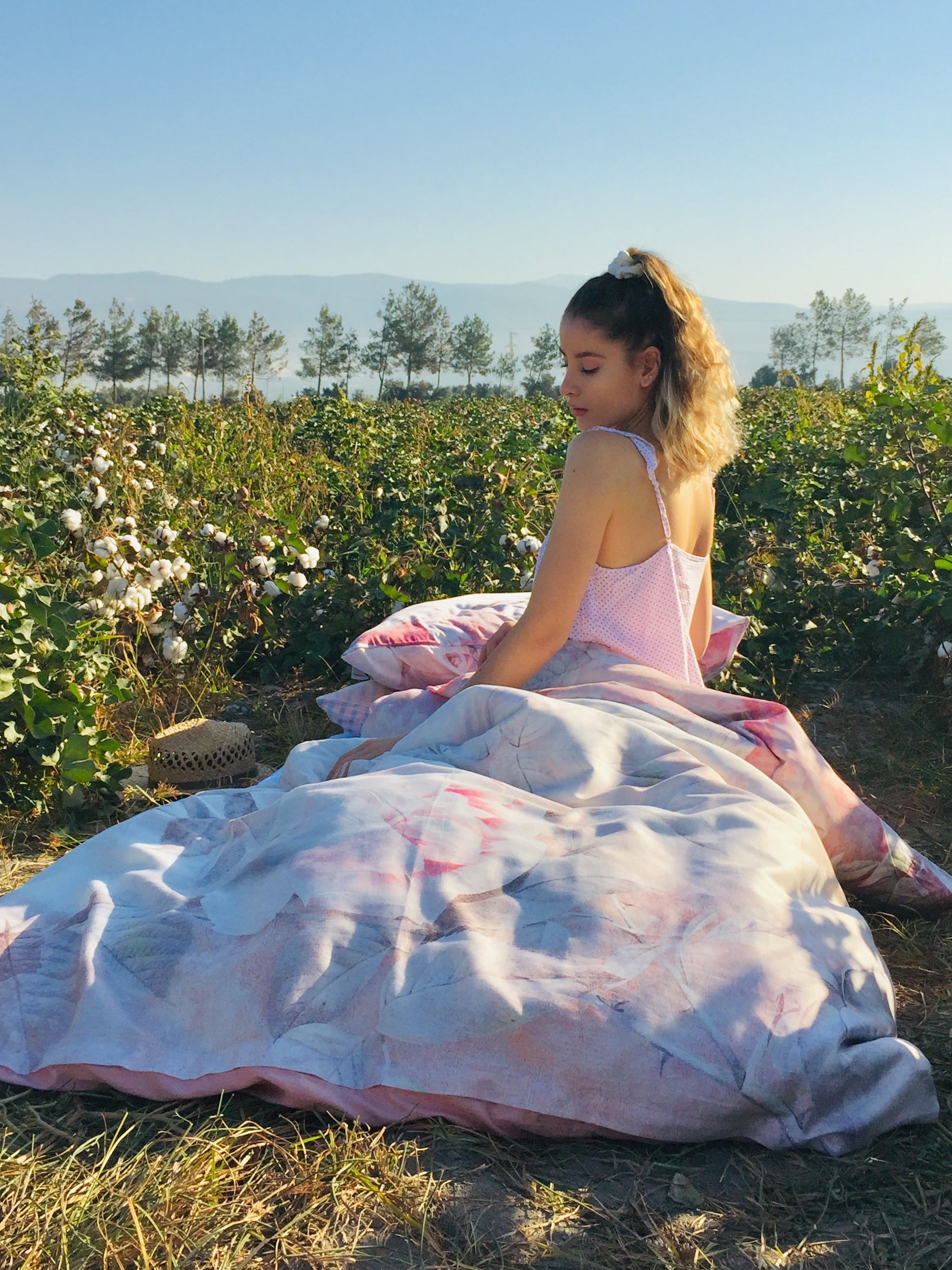 Eine junge Frau mit lockigem, schulterlangem Haar sitzt in einem Baumwollfeld, eingehüllt in eine Bettdecke mit einem floralen Muster in sanften Rosa- und Beigetönen. Sie trägt ein zartes, rosa gepunktetes Trägerkleid und blickt zur Seite. Neben ihr liegt ein geflochtener Strohhut auf dem Boden. Im Hintergrund sind Baumwollpflanzen mit weißen Blüten, eine Baumreihe und entfernte Berge unter einem klaren blauen Himmel zu sehen. 