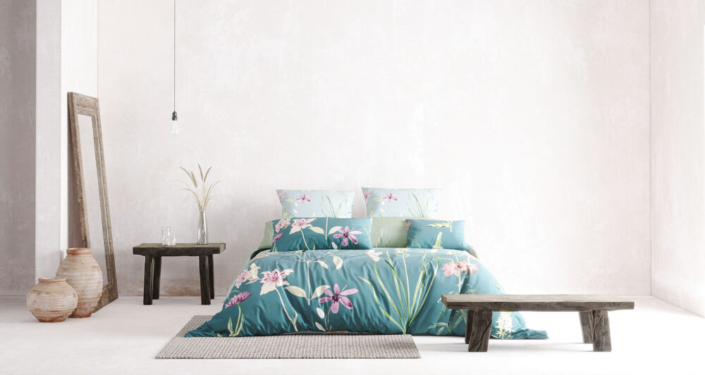 Ein minimalistisch eingerichtetes Schlafzimmer mit einer türkisfarbenen Bettwäsche, die mit zarten rosa und lila Blumen sowie grünen Blättern verziert ist. Das Bett steht vor einer hellen, strukturierten Wand. Links daneben lehnt ein rustikaler Holzrahmen-Spiegel an der Wand, daneben stehen zwei Keramikvasen auf dem Boden. Ein kleiner Holztisch mit einer Vase und zwei Gläsern befindet sich ebenfalls links vom Bett. Eine einzelne, minimalistische Hängelampe sorgt für dezente Beleuchtung. 