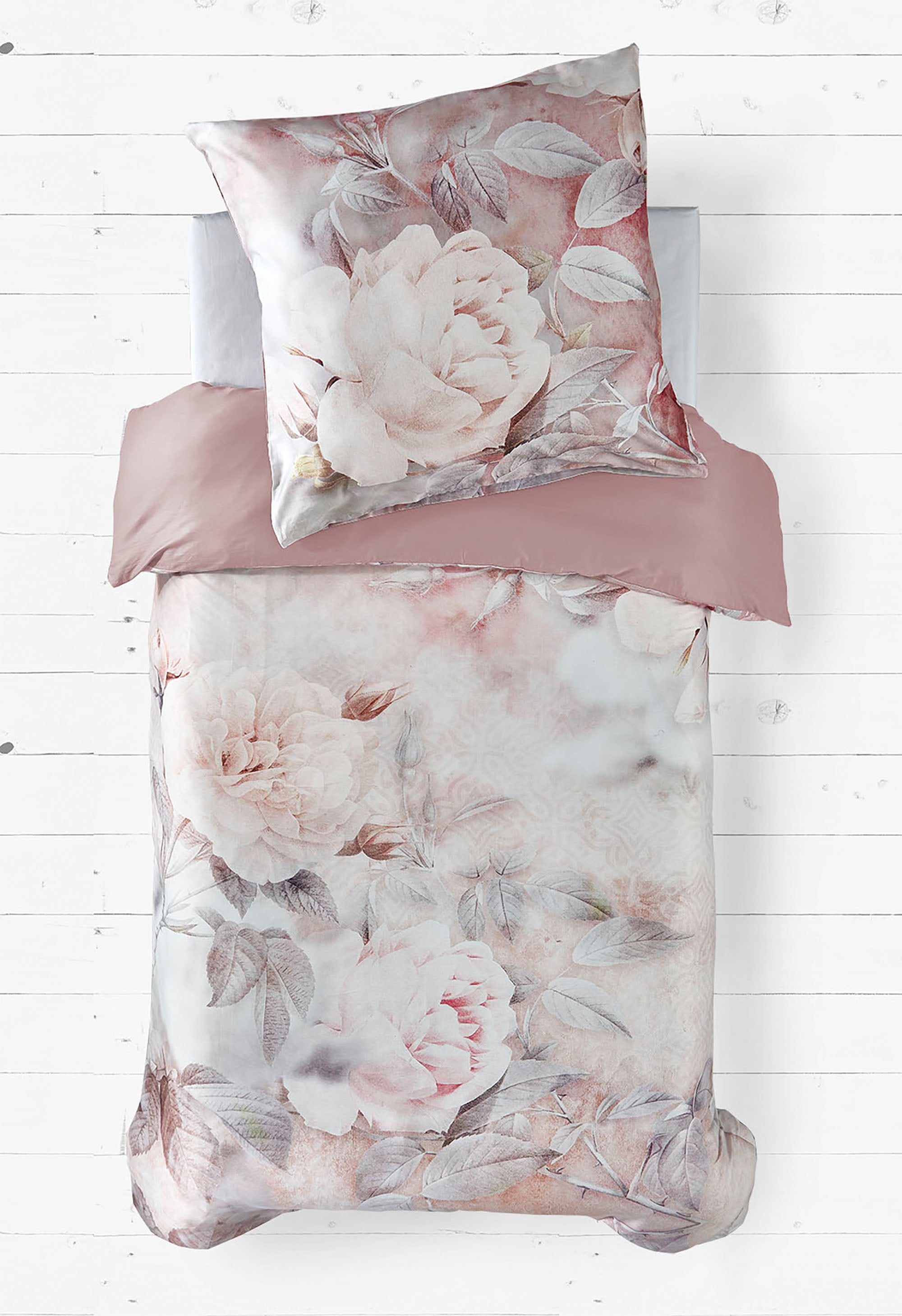 Das Bild zeigt ein Bettwäsche-Set mit einem romantischen floralen Design. Der Bettbezug und der Kissenbezug sind mit großen, zarten Rosen in sanften Rosa- und Beigetönen verziert, ergänzt durch blasse Blätter in Grau- und Taupe-Tönen. Die Rückseite des Bettbezugs scheint in einem einfarbigen, warmen Altrosa gehalten zu sein. Die Bettwäsche liegt auf einem weißen Holzboden, was einen eleganten und harmonischen Kontrast bildet.